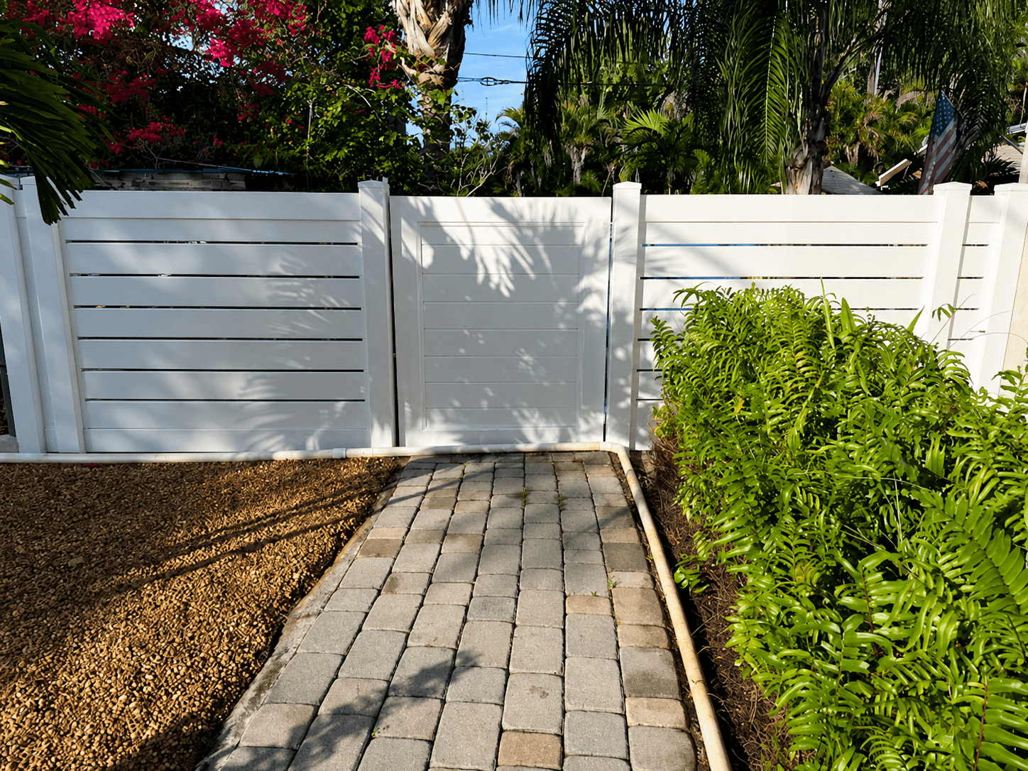 White vinyl horizontal-slat fence and gate with paver walkway and tropical landscaping